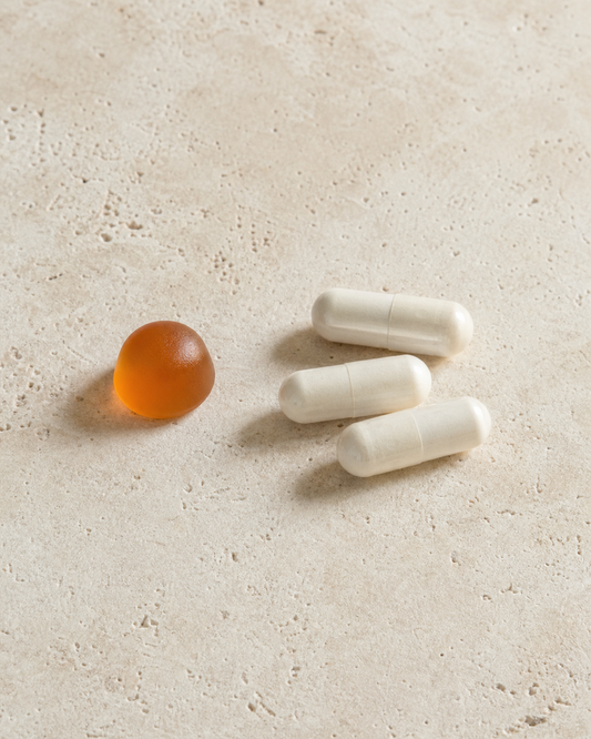 One small amber-orange dome gummy on the left and three plain off-white two-piece capsules on the right of an off-white travertine surface in soft side daylight. No bottle, text, paper, person, or additional props.